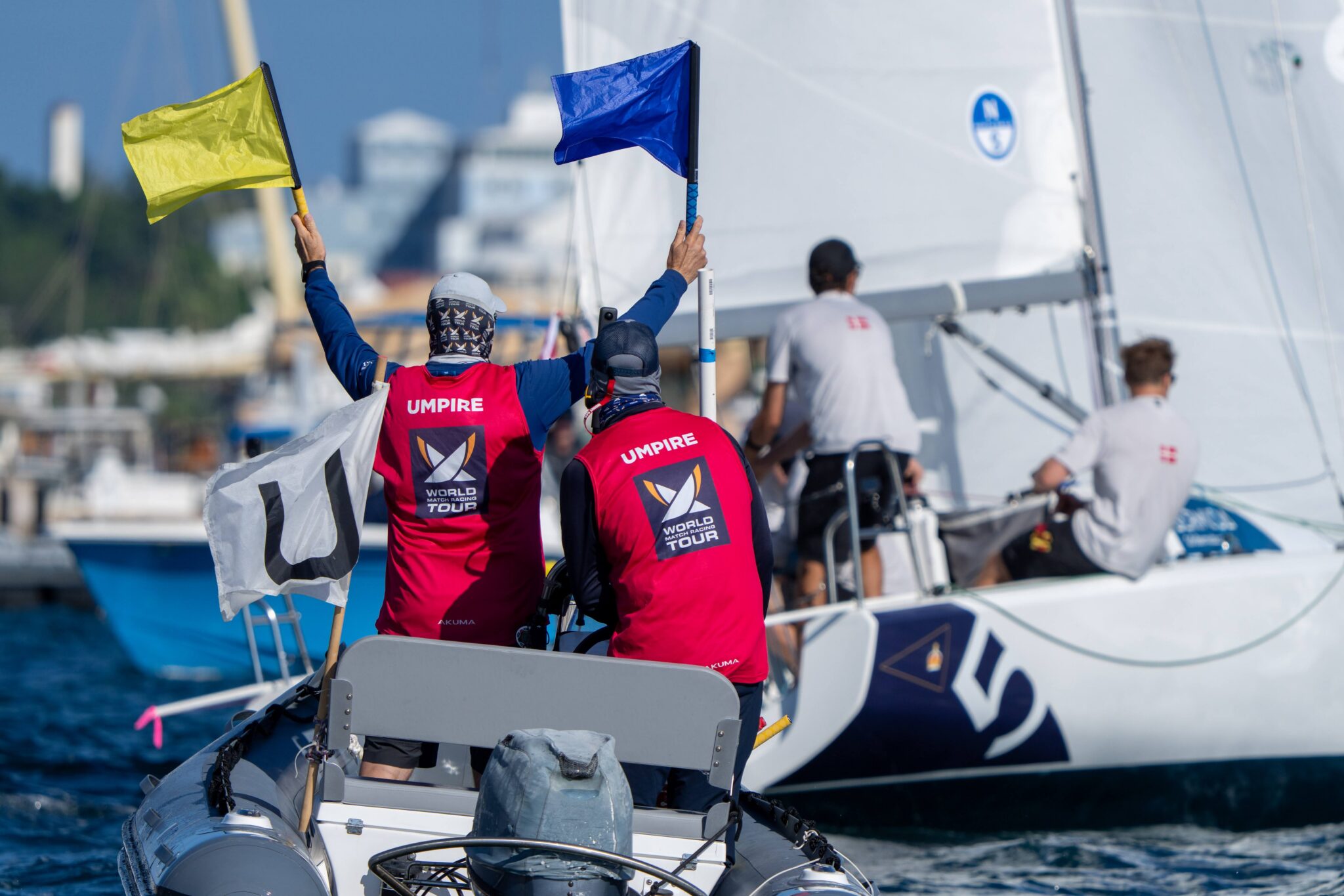 Dwóch sędziów World Match Racing Tour z żółtą i niebieską flagą na pontonie obok jachtu